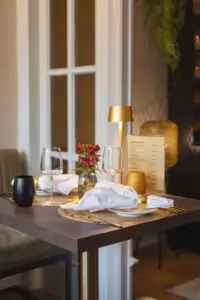 Gedeckter Restauranttisch am Fenster mit goldener Tischlampe, Weingläsern und roten Blumen